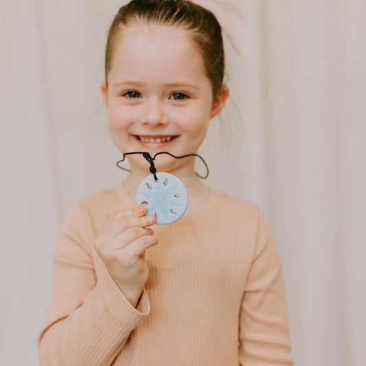 Young girl holding a blue snowflake chewable pendant against a plain background