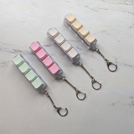 Keyboard fidget clicker keychains in green, pink, cream and beige on a light background