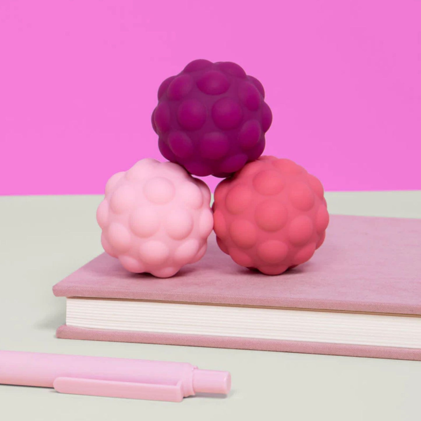Three Speks Odds pink silicone fidget magnets on a pink notebook with a pink pen, against a pink background