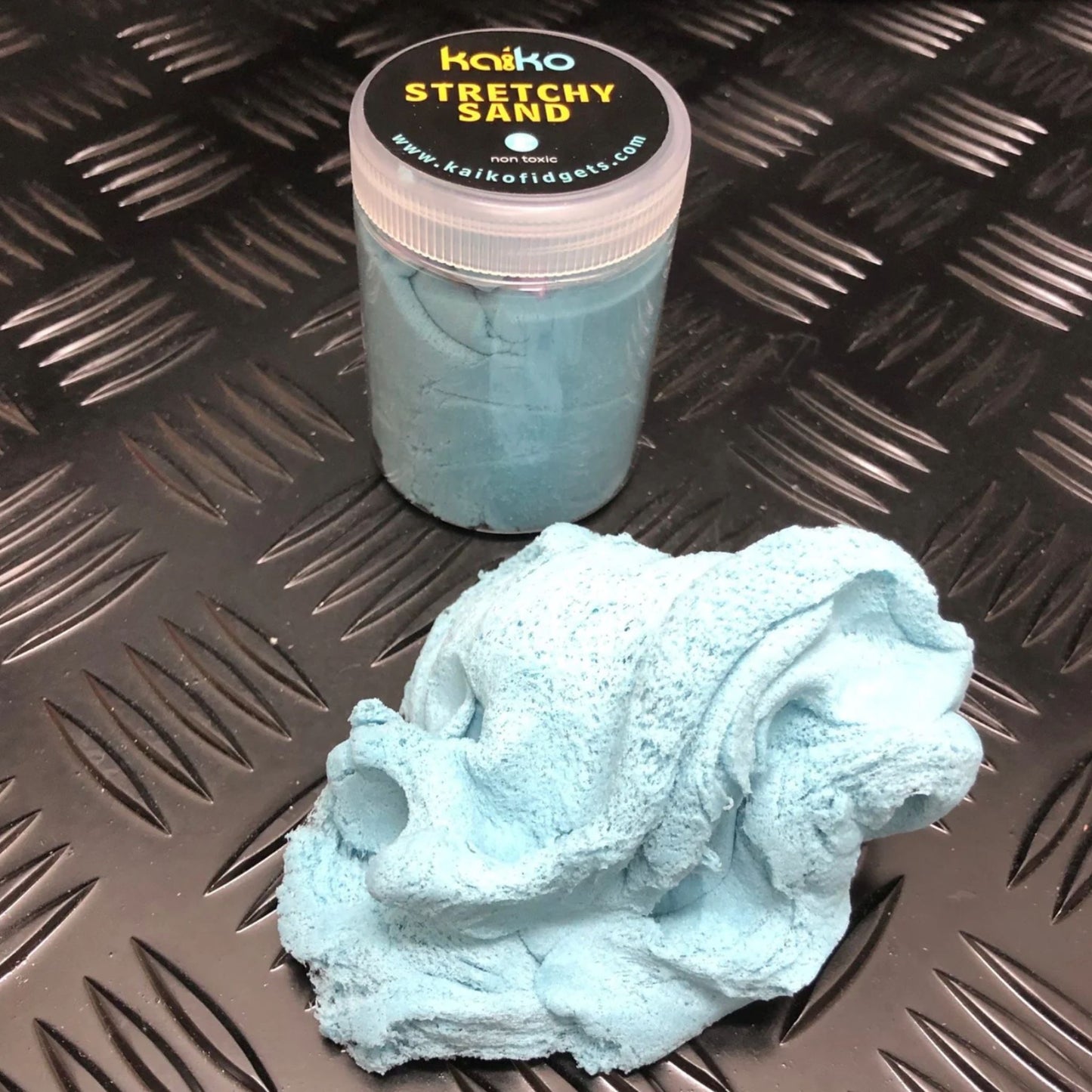 Blue stretchy sand with a container labeled 'Kakko Stretchy Sand' on a textured black surface.