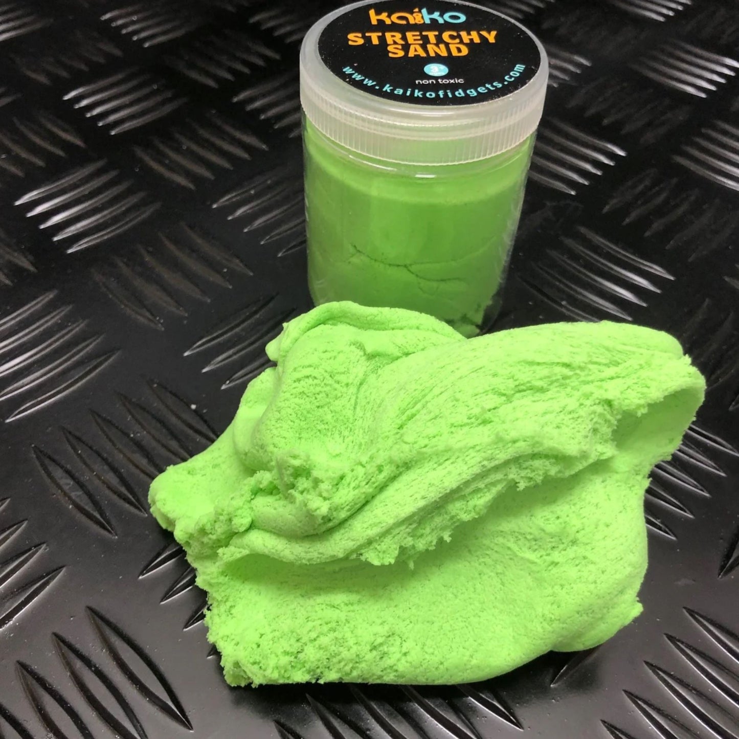 Green stretchy sand with a container labeled 'Kaiiko Stretchy Sand' on a textured black surface.