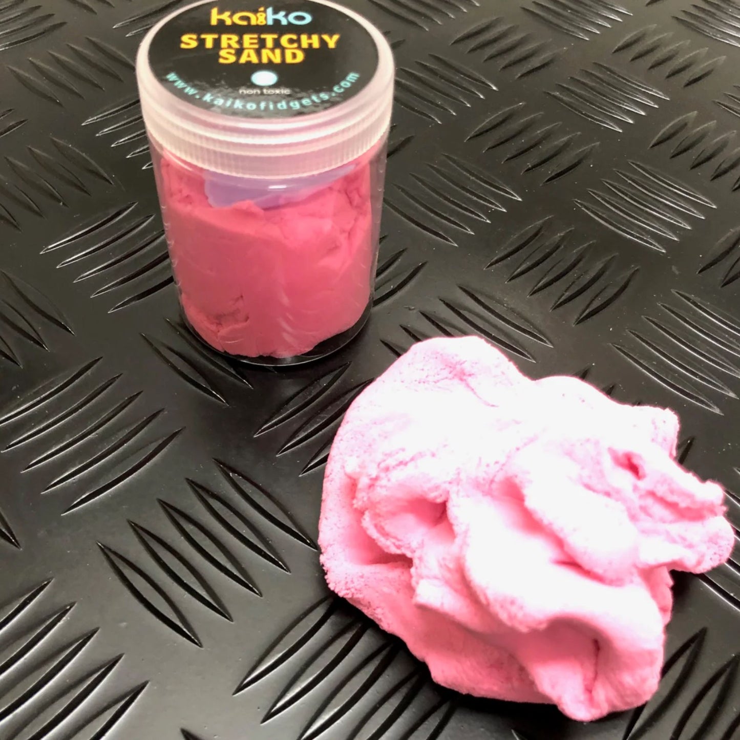 Pink stretchy sand with a container labeled 'Kaiiko Stretchy Sand' on a textured black surface.