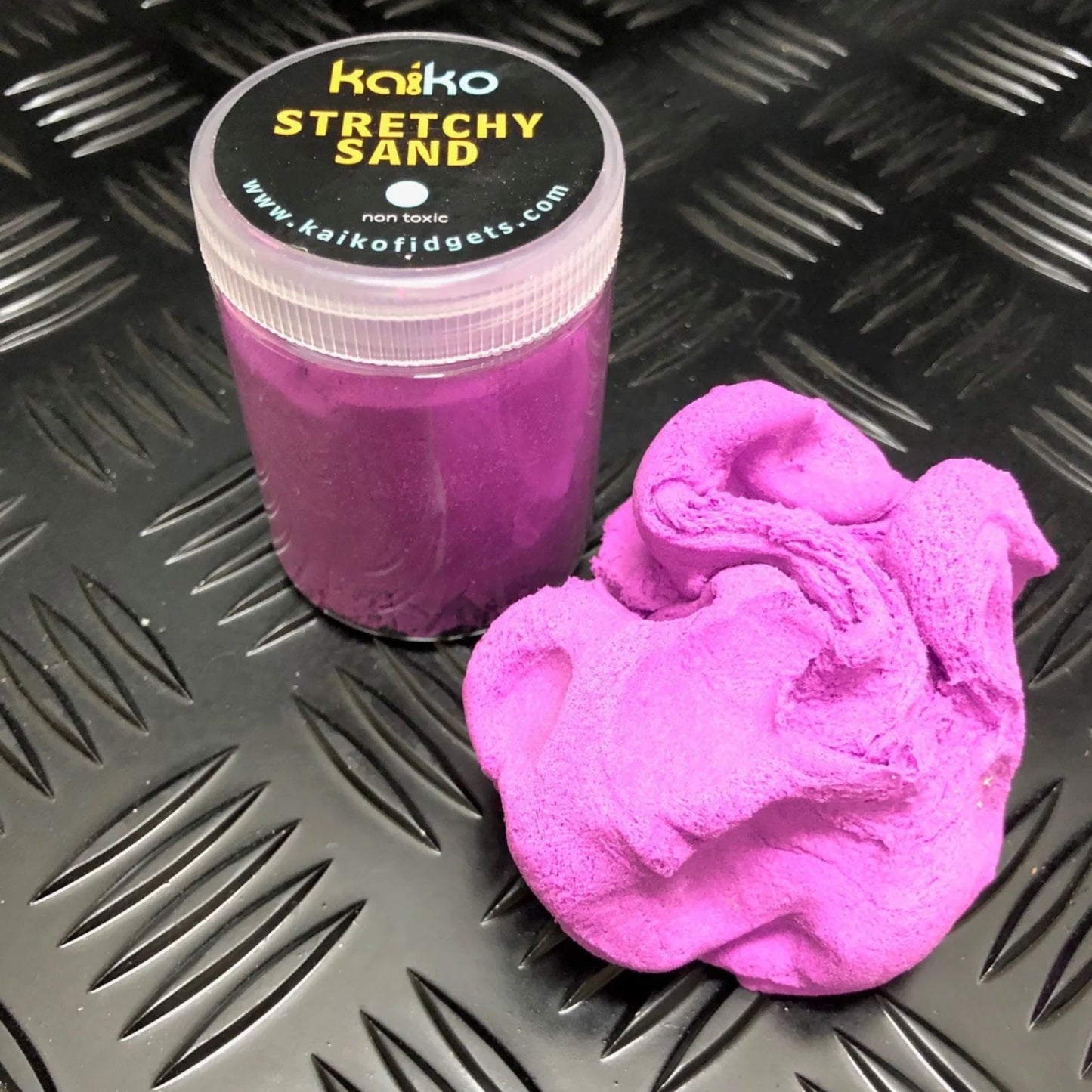 Purple stretchy sand with a container labeled 'Kaiko Stretchy Sand' on a textured black surface.