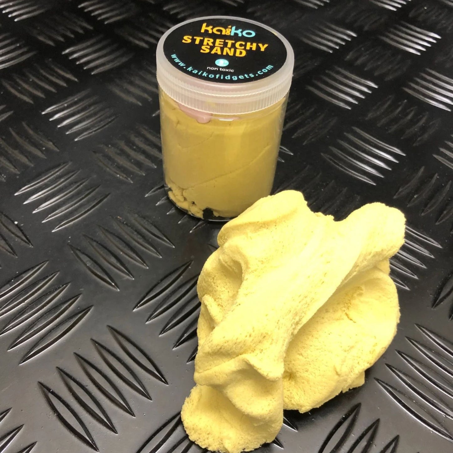 Yellow stretchy sand with a container labeled 'Kwikko Stretchy Sand' on a textured black surface.
