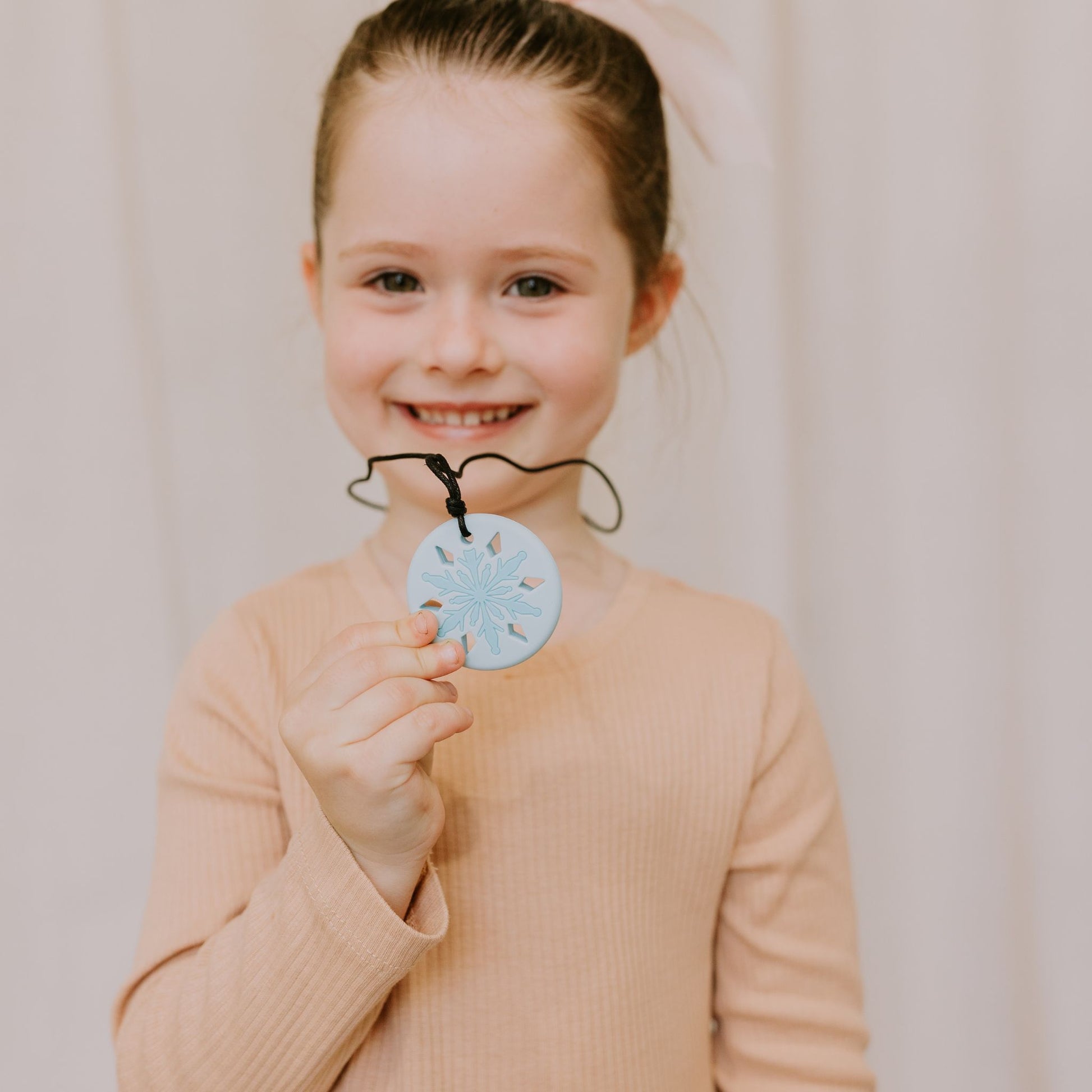 Young girl holding a blue snowflake chewable pendant against a plain background